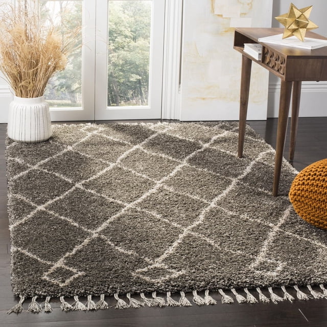 Safavieh Moroccan Fringe Lavern Geometric Shag Area Rug in grey with white diamond pattern and fringed edges, placed on dark hardwood floor beside a wooden side table and a white vase with dried grasses, ideal for modern or country-chic accent decor.