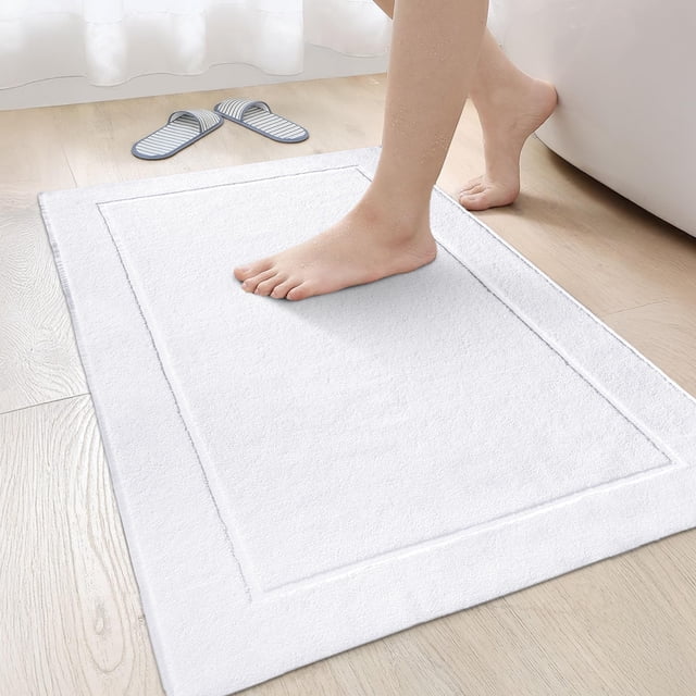 Canadian Linen Yukon Bath Mats, 2 Pack, 20"x34", lightweight quick-dry soft absorbent cotton bathroom mats in white with a picture frame design. A foot is stepping on the mat, highlighting its soft terry texture and absorbency. Ideal for bathroom rug sets, these mats are placed on a wooden floor near a bathtub, with striped slippers visible in the background. Suitable for use in bathrooms, near showers, or as multipurpose floor mats.
