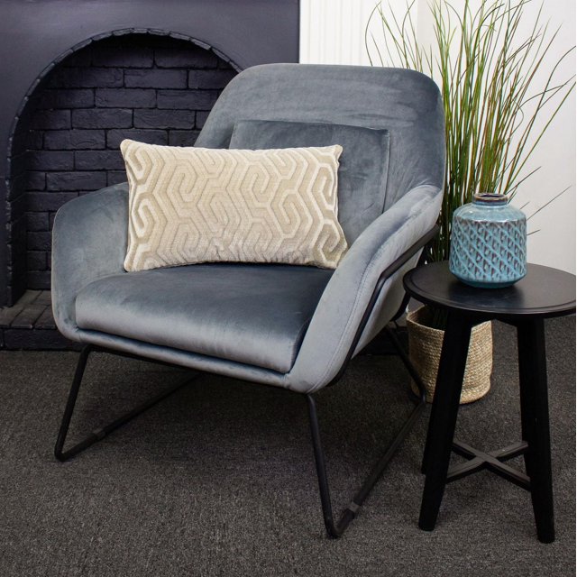 Coussin de Luxe Zulu Rempli de Plumes avec Motif Géométrique beige sur un Fauteuil en Velours Gris avec Pattes en Métal Noires, à Côté d'Une table d'Appoint Ronde Noire Tenant un vase en Céramique Texturé Bleu et un Panier Tressé avec une Haute Herbe Verte, Adossé à une Cheminée en Briques Noires et un Mur Blanc.