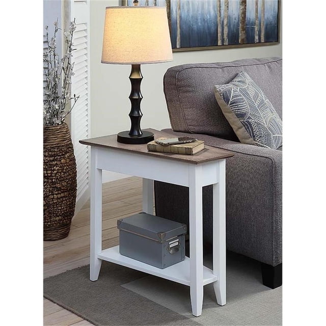 Convenience Concepts American Heritage Table d'Appoint en Coin, Fini Bois Blanc, Présentant un design Triangulaire avec un Plateau Spacieux et une Tablette Inférieure pour un Rangement Supplémentaire. Fabriquée en Bois Massif et Manufacturé avec une Finition Blanche, Elle Est Présentée Placée à Côté d'Un sofa Gris dans un Décor de Salon. la Table Est Conçue avec une Lampe Noire, des Livres et une Boîte Décorative sur l'Étagère Inférieure.