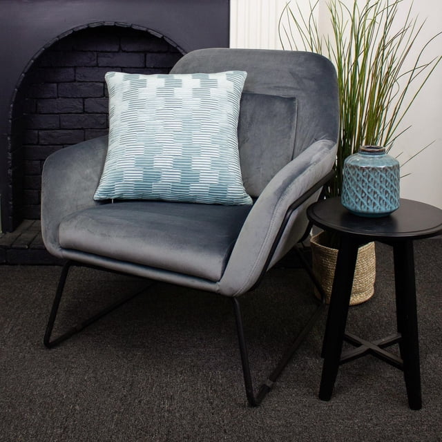 Coussin de Luxe Treble avec un Motif Géométrique Doux Bleu et Blanc sur un Fauteuil en Velours Gris avec des Pattes en Métal Noir. le Coussin Carré de 18 Pouces Rehausse un Salon Moderne avec une table d'Appoint Noire Tenant un vase en Céramique Bleu et une Haute Herbe Verte dans un Panier Tressé Près d'Un Foyer en Briques Noires. Utilisé Souvent comme accent Décoratif Ou pour un Confort d'Assise Accru.