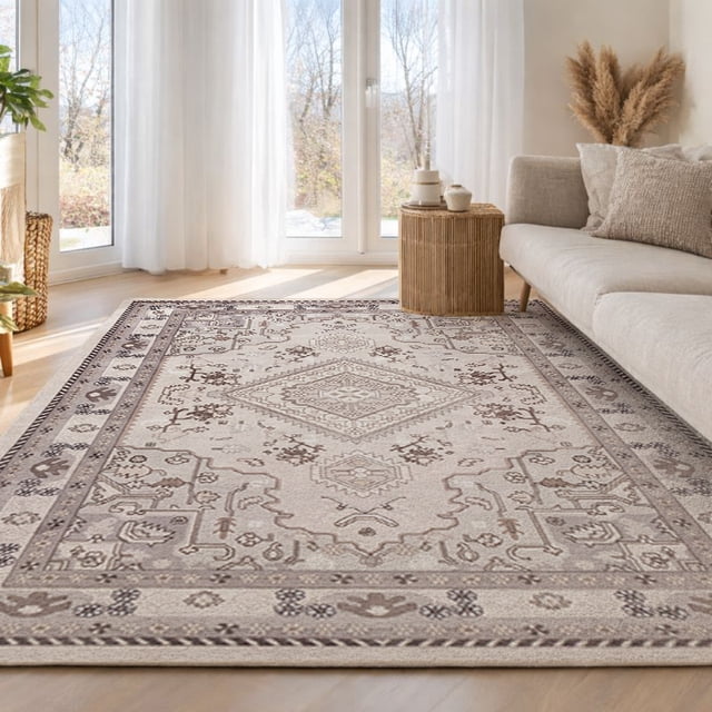 ECARPET Oriana Machine Washable Traditional Area Rug in muted beige and taupe tones with a modern Heriz-inspired geometric pattern and softly distressed finish, featuring anti-slip backing and machine-washable convenience, displayed on a light hardwood floor in a bright living room with sheer curtains, a beige sofa with textured cushions, a wicker side table, and dried pampas grass, suitable for living rooms, bedrooms, or hallways.