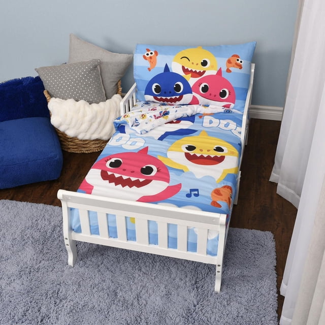 Baby Shark 3 Piece Toddler Bedding Set with reversible comforter, fitted sheet, and pillowcase on a white toddler bed. The comforter features large colorful Baby Shark characters on a light blue striped background with musical notes and small orange fish. The fitted sheet and pillowcase have matching Baby Shark prints. The set fits a standard crib mattress and is made of soft polyester. The bed is on a gray shag rug with a blue plush chair and a basket holding gray and white pillows nearby.