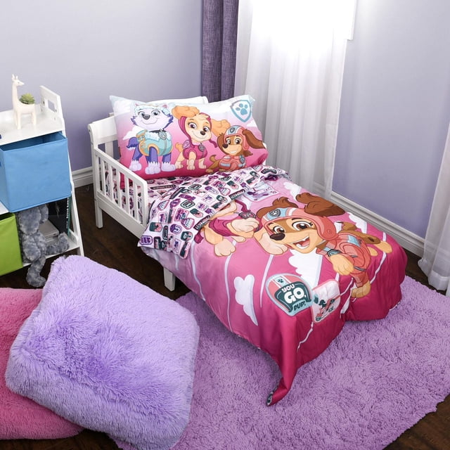 Paw Patrol Skye 3-Piece Toddler Bedding Set on a white toddler bed with a reversible comforter featuring bright pink and purple colors and high-definition prints of Skye, Liberty, and Everest. The fitted sheet shows a pattern of Paw Patrol badges. The set is in a toddler room with lavender walls, sheer white curtains, a lavender plush rug, and soft cushions on a dark hardwood floor.