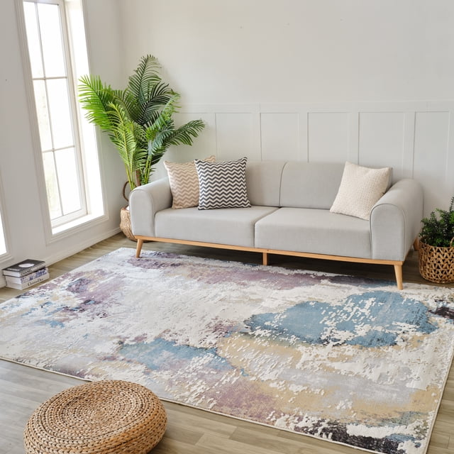 Modern abstract beige and blue indoor area rug placed in front of a light gray sofa with chevron and textured pillows, styled in a bright living room setting with plants.