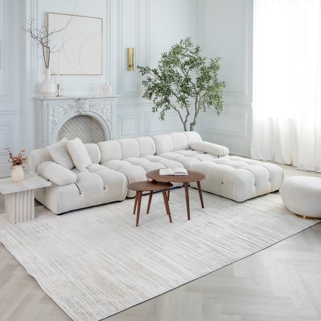 Rug Branch Astra Modern Indoor Area Rug in beige cream with abstract pattern, placed in a bright living room featuring a tufted cream sectional sofa, two nested wooden coffee tables with an open book, a round ottoman, and a large green potted plant near a white ornate fireplace and tall windows with sheer curtains.