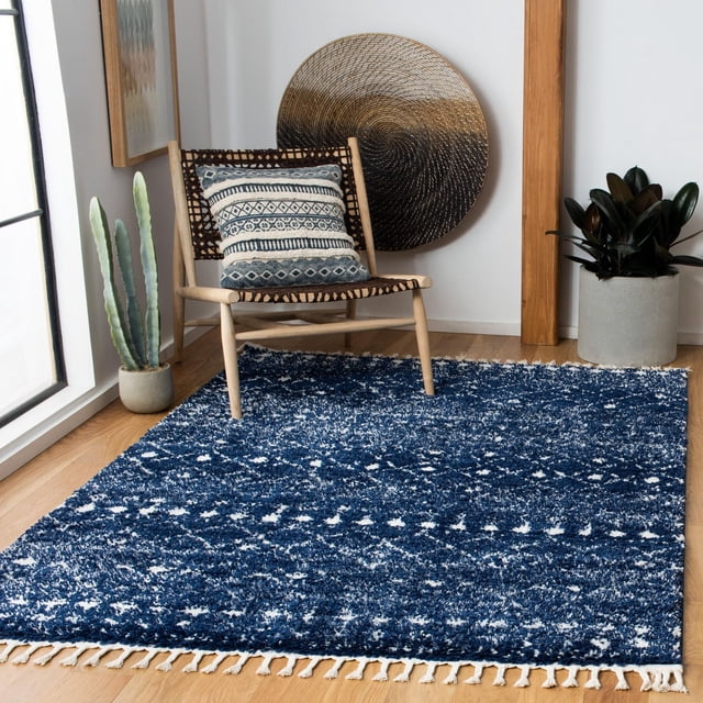 Safavieh Berber Fringe Robynne Aztec Shag Area Rug in deep blue with white Aztec tribal patterns and 1.25-inch pile, featuring fringed edges on a light wood floor, styled with a woven chair with a patterned cushion, a cactus plant, round woven wall art, and a potted leafy plant, suitable for adding a bohemian accent to living spaces.