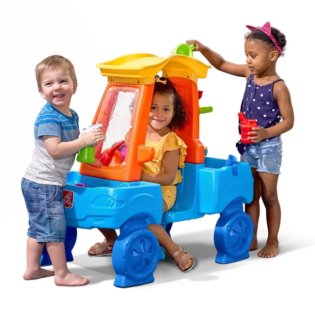 Step2 Centre d'Éclaboussures pour Lavage de Voiture Est une table d'Eau en Plastique Bleu et orange en Forme de Voiture avec un Toit Jaune, une Fenêtre avant Transparente et un Essuie-Glace Rouge; Trois Enfants Jouent avec des Bouteilles Vaporisateurs et des Gobelets d'Eau, Favorisant le Jeu Extérieur social et Sensoriel.