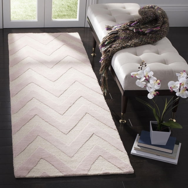 Safavieh Cambridge Kaitlyn Zig Zag Stripes Area Rug with hand-tufted wool and textured zig zag stripes in cream and light pink, placed on dark hardwood floor beside a beige tufted bench with brass legs, a multicolored throw, and a white orchid on books, suitable as a durable accent rug for home or office.