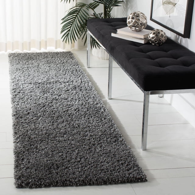 Gray SAFAVIEH August Carlene solid plush shag area rug runner placed on white wooden floor beside a modern black tufted bench with chrome legs and decorative metallic spheres.