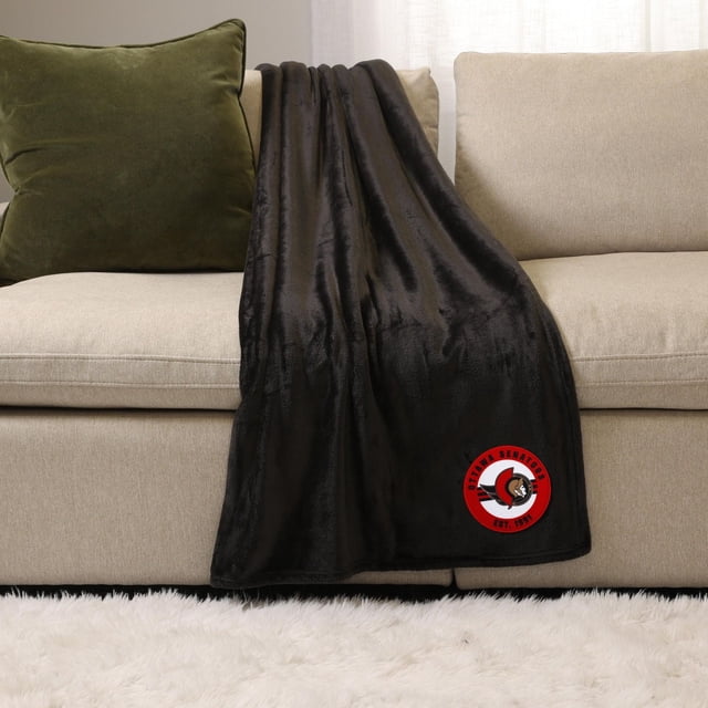 NHL, NBA, MLB Black Sports Throw Blanket, 50" x 60", draped over a beige sofa with a green cushion, made of soft plush 100% polyester fabric featuring an Ottawa Senators logo patch in red and black near the bottom edge, ideal for cozy indoor use and extra warmth.