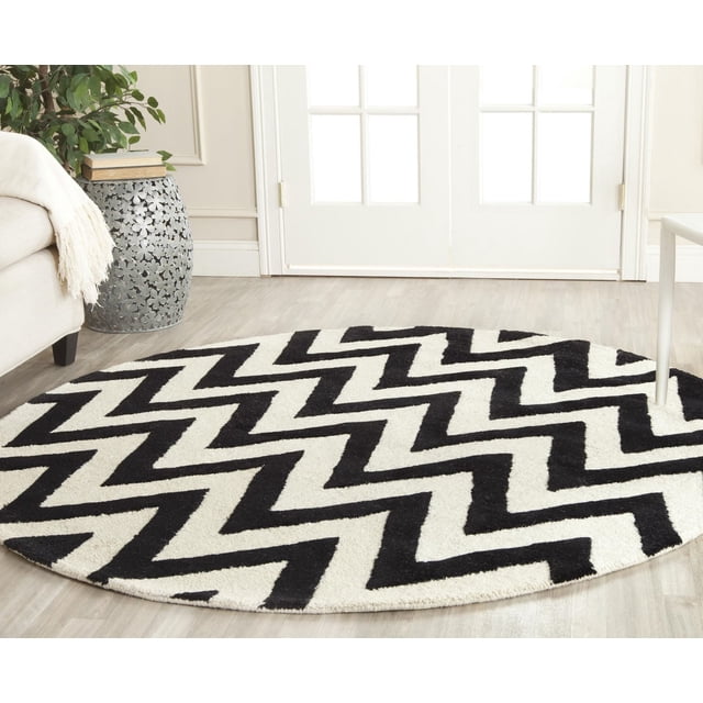 Safavieh Cambridge Kaitlyn Zig Zag Stripes Area Rug, a round hand-tufted wool rug with a black and white zigzag pattern, placed on light wood flooring in a bright room with white French doors and neutral furniture, ideal for adding modern American style to home or office decor.