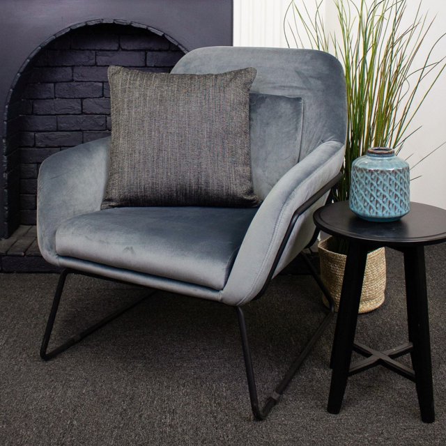 Remy Luxury Cushion, a decorative throw pillow in textured dark grey fabric, placed on a modern grey velvet armchair with black metal legs, beside a black round side table holding a blue ceramic vase and tall green grass in a woven basket, set against a black brick fireplace and white paneled wall, suitable for enhancing home décor and comfort.