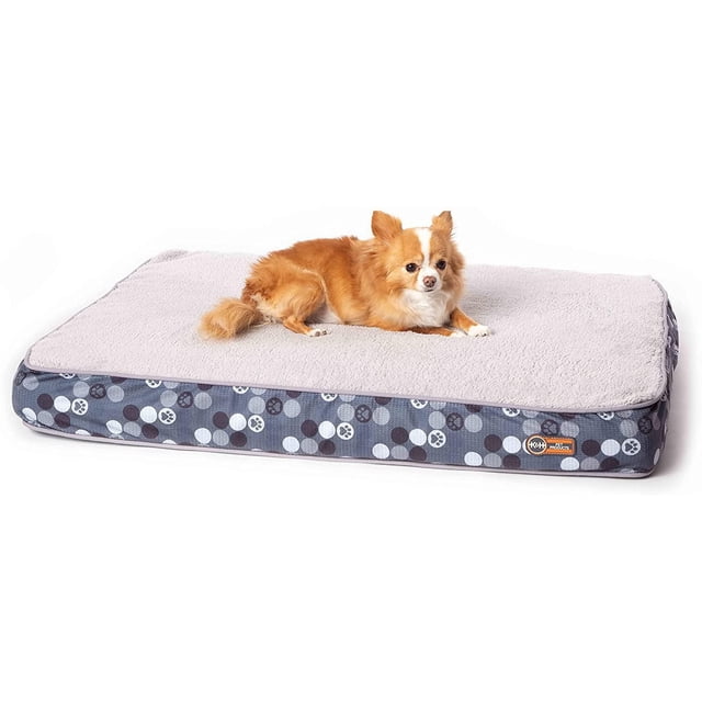 K&H Pet Products Superior Orthopedic Dog Bed in grey with a small tan and white dog resting on the soft fleece side. The 27 by 36 inch rectangular bed features a reversible cover with paw print patterns and high-density orthopedic foam for joint and bone support, ideal for pets needing extra comfort and stability.