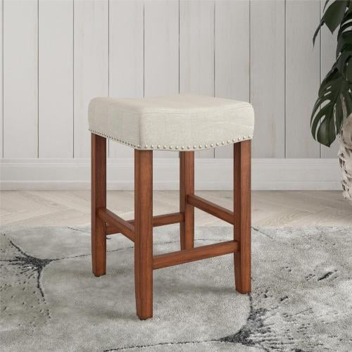 Upholstered Counter Height Bar Stool featuring a 24-inch height farmhouse-inspired design with a slightly curved foam-cushioned saddle seat in beige linen upholstery. The seat is accented with brass nailhead trim and supported by strong rubberwood legs with side footrests and capped feet to protect floors. Ideal for kitchen islands or high-top dining tables, it includes all parts, tools, and instructions for easy assembly.