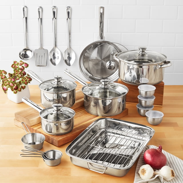 Mainstays Stainless Steel Cookware Set, 24-piece displayed on a wooden countertop with white subway tile backsplash, featuring saucepans, Dutch oven, fry pan, casserole with rack, ramekin bowls with lids, metal measuring cups, and hanging stainless steel utensils, all with tri-ply base for even heating and compatible with induction, gas, and electric stovetops.