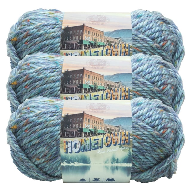 Lion Brand Hometown Yarn - Blue Sky, three skeins of thick, machine-washable blue tweed yarn with orange and yellow flecks, wrapped with labels featuring a small town and mountain scene, ideal for fast knit and crochet projects and suitable for school or sports team crafts.