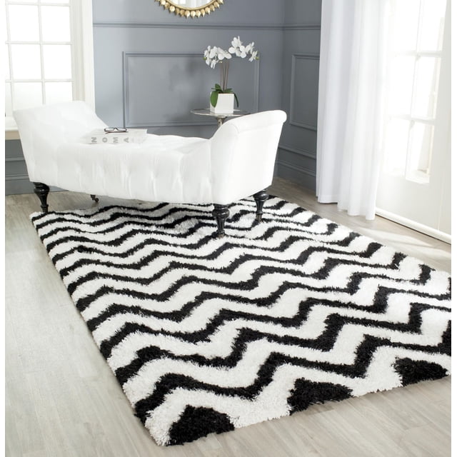 Safavieh Barcelona Carmella Zigzag Stripes Shag Area Rug featuring a black and white zigzag pattern with plush shag texture on light wood flooring, placed under a white tufted chaise lounge with black legs in a bright room with gray paneled walls and white curtains, suitable for modern accent rugs and cozy floor décor.