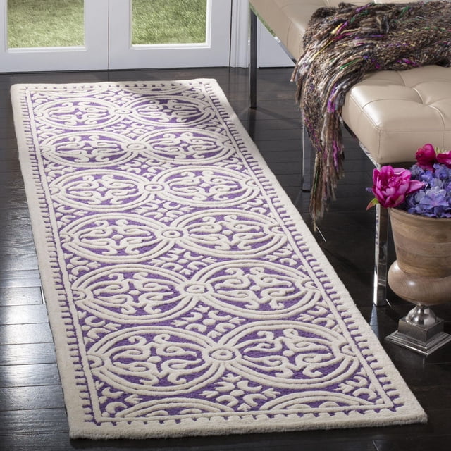 SAFAVIEH Cambridge Leslie Geometric Area Rug featuring hand-tufted wool with a purple base and cream geometric cut and loop pile patterns, displayed on dark hardwood flooring beside a beige leather bench with a textured throw and a wooden vase with pink and purple flowers, ideal for adding classic American style and texture to home or office decor.