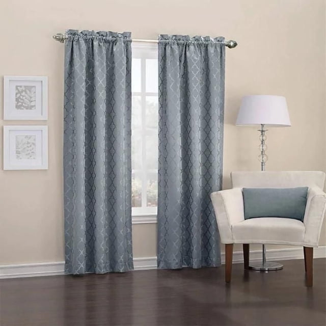 Sun Blocker Daniella Thermal Rod Pocket Curtain in blue with embroidered geometric pattern, featuring thermal backing that saves energy, blocks light, and reduces noise, hanging on a silver rod in a beige room with dark hardwood floors, next to a cream armchair with a blue pillow and a silver floor lamp.