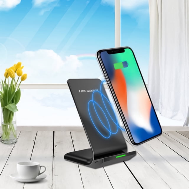 TopLLC Fast Wireless Charger, Certified, 15W Max Wireless Charging Stand compatible with Android and iOS. The black stand features a sleek, vertical design with two coils for faster charging and a wider charging area. A smartphone is shown charging on the stand, positioned in a bright, modern room with a wooden floor and a window view. The charger includes a cooling hole for heat dissipation and supports charging through phone cases. Package includes a wireless charger, manual, and data cable.