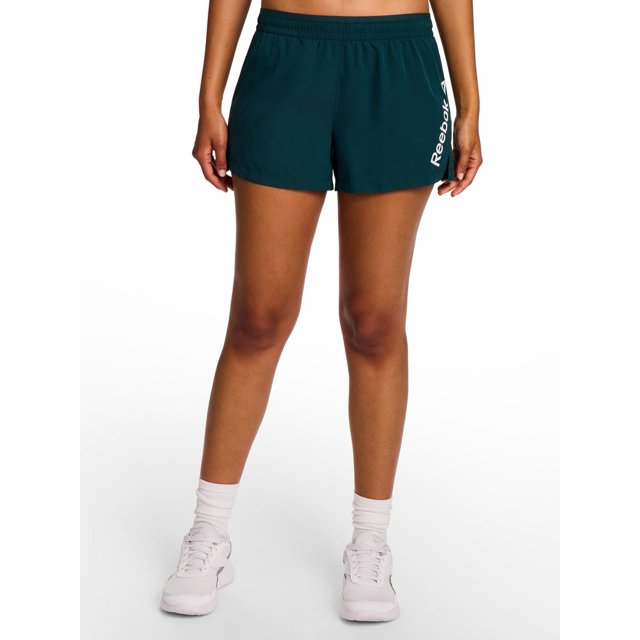 Reebok Women's Staple Running Short with Mesh Lining in dark teal, front view on a model from waist to mid-thigh, made of lightweight polyester with elastic waistband and drawcord, side slits, built-in mesh liner, and white Reebok logo on left leg, suitable for running or training, shown with white socks and sneakers on white background.