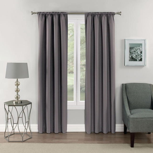 Eclipse Samara 63" rod pocket blackout panel in dark grey with textured tone-on-tone design hanging on a silver curtain rod over a white-framed window in a modern living room with light grey walls, a grey upholstered chair, a silver lamp on a geometric metal side table, and a framed black-and-white photo. These thermal curtains block over 99% of light, reduce noise, and save energy.