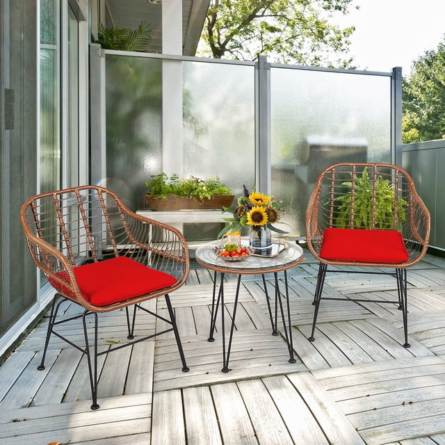 Costway Ensemble Bistro en Rotin pour Patio 3 Pcs Table Basse Fauteuil Jardin Coussin Rouge. l'Ensemble Comprend Deux Fauteuils en Rotin Bruns avec Coussins Rouges et une Table Ronde à Dessus en Verre Trempé. les Chaises Présentent un design Tressé avec des Pieds en Fer Noir, et la Table a un Cadre Robuste avec des Patins Antidérapants. l'Ensemble Est Exposé sur un Patio en Bois avec Verdure et un Écran d'Intimité en Verre Givré en Arrière-Plan, Idéal pour les Espaces Extérieurs comme les patios, Jardins Ou Balcons.