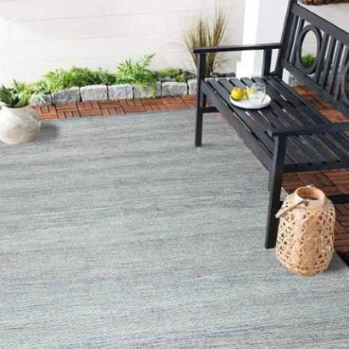 Tapis Océan 8'x10 Pieds - Tapis d'Extérieur Bleu Naturel en Fil RPET Recyclé à texture Tissée dans des Tons Bleu Pâle et Gris, Posé sur un patio avec un Banc en Bois Noir Tenant un Plateau avec un Verre et Citron, Plantes en Pot, et une Lanterne Tissée sur un Plancher de Terrasse en Bois.
