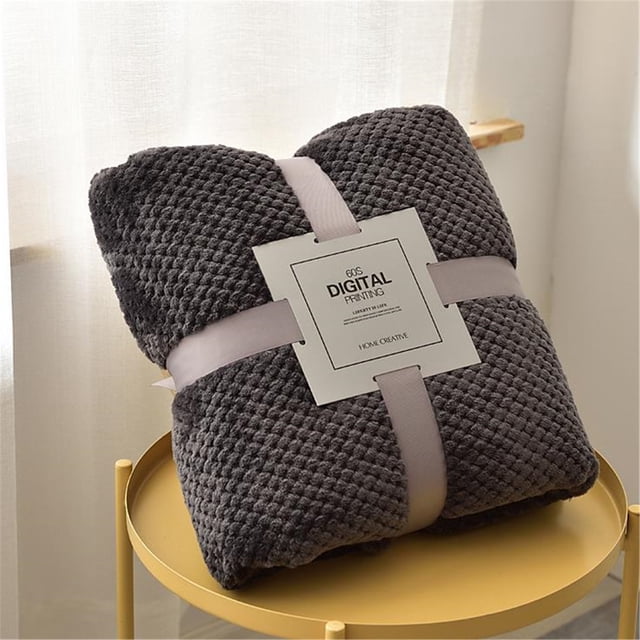 Couverture de Sofa Unie Anti-Rayures de Chat en Flanelle Peluche, Montrée en Couleur Gris Foncé. la Couverture Est Soigneusement Pliée et Fixée avec un Ruban Gris Pâle, avec une Étiquette Indiquant « Impression Numérique des Années 60 ». le Tissu en Molleton Corail Texturé Est Doux, Respirant et Chaud, Idéal pour une Utilisation Intérieure et Extérieure. la Couverture Mesure 100x150 cm et Est Lavable à la machine. Exposée sur une table Ronde Jaune dans une Pièce Légèrement Éclairée.