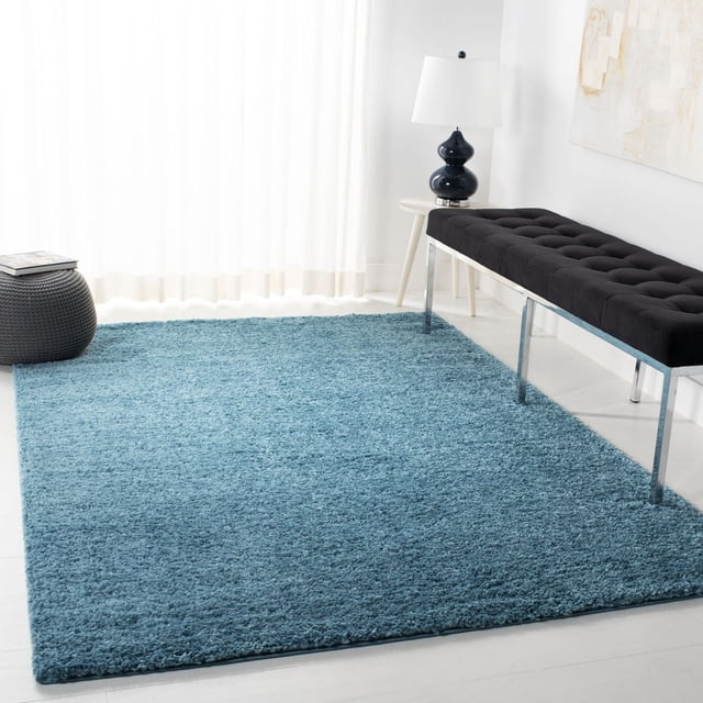 SAFAVIEH August Carlene Tapis Shag en Peluche Uni Bleu sur un Plancher Lumineux dans une Pièce Lumineuse avec des Rideaux Blancs Transparents, un Banc Capitonné Noir avec des Pattes Chromées, une table d'Appoint Blanche et une Lampe en Céramique Noire avec un Abat-Jour Blanc, Idéal pour Ajouter Douceur et Confort aux Espaces de Vie.