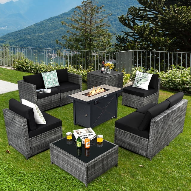 Gymax 9 PCS Patio Rattan Furniture Set Fire Pit Table Storage Grey W/ Cover Black. This outdoor patio conversation set includes four armless sofas, two corner sofas, a coffee table with a tempered glass top, a storage box, and a 42-inch fire pit table with a 60,000 BTU propane heat output. The furniture is crafted from durable PE rattan and a powder-coated steel frame, featuring black cushions with detachable covers for easy cleaning. The fire pit table has a ceramic top and comes with a protective cover and lava rocks. Suitable for gardens, patios, or backyards, the set offers versatile configurations for gatherings. The background shows a lush green lawn with a scenic mountain view.