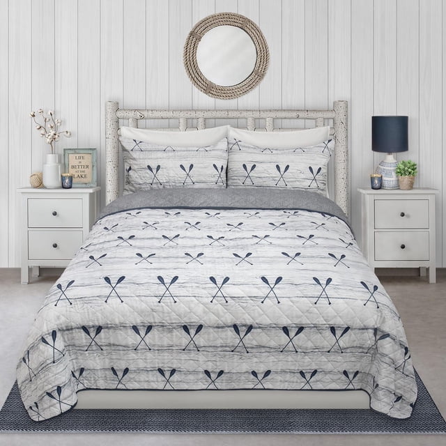 Safdie & Co. Quilt 3PC Set K Paddles on a bed with a rustic wooden headboard in a coastal-style bedroom. The quilt and two shams feature a white and light gray background with navy blue crossed paddle prints and diamond quilting. Made of soft, breathable 100% polyester microfiber, this set resists fading, stains, shrinking, wrinkles, dust mites, and bacteria. The room includes white paneled walls, a round rope mirror, white nightstands with a navy lamp, plants, candles, and a framed sign reading 'Life is better at the lake'. This quilt set adds subtle nautical decor and comfort to any bedroom.