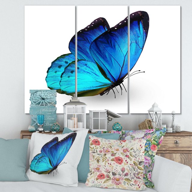 Designart Vibrant Blue Butterfly Canvas Wall Art featuring a large blue butterfly across three gallery-wrapped cotton canvas panels with a white background, displayed above a wooden console with decorative lanterns and floral cushions, suitable for modern home décor and wall art.