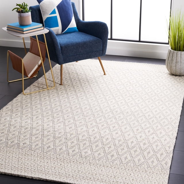 SAFAVIEH Marbella Ema Geometric Area Rug featuring a hand woven flat weave design with subtle diamond patterns in neutral tones, placed on dark hardwood floors under a blue upholstered armchair with a geometric pillow, beside a gold frame side table with books and a small plant, near a tall green plant by a large window, suitable for durable, cushioned accent rug use in home interiors.