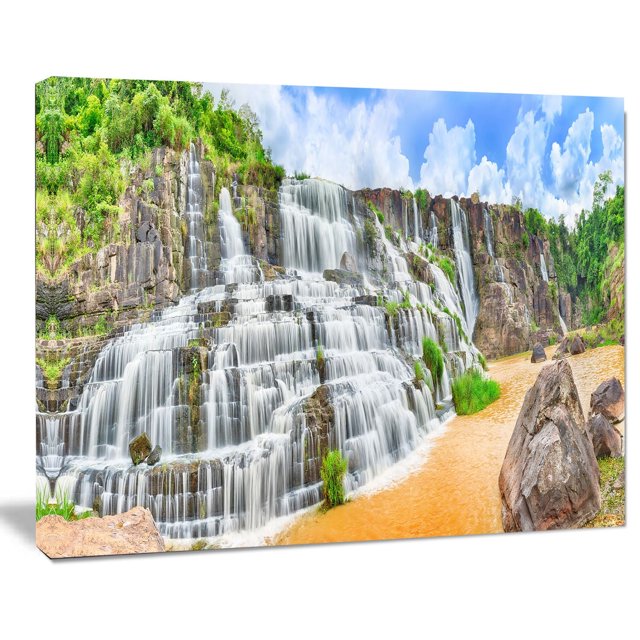 Design Art Pongour Waterfall Canvas Print featuring a gallery wrapped cotton canvas with a high-resolution image of a cascading multi-tiered waterfall over rocky cliffs with green vegetation, flowing into a muddy river under a blue sky with clouds, ready to hang with included hardware, suitable for enhancing home or office wall décor.