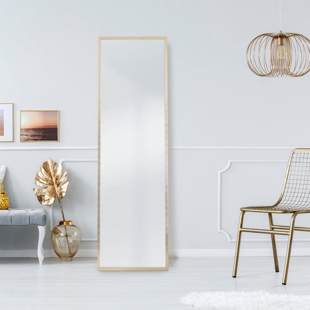 Home Trends 17''*58'' Easel Mirror, Champagne with a slim champagne frame featuring subtle linear patterns, standing in a bright room with white walls and light wood flooring, flanked by a grey bench with gold accents and a gold wire chair beneath a gold pendant light, ideal for full-length reflections and enhancing bedroom or living space decor.