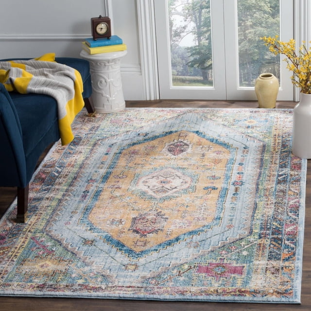 SAFAVIEH Bristol Leola Traditional Area Rug with a distressed antique-style hexagonal medallion pattern in muted yellow, blue, and pastel tones, made of polyester fibers, placed on a hardwood floor in a bright room with a blue sofa, yellow and gray throw, white ceramic side table with books and clock, and vases with yellow flowers near glass doors, suitable for adding vintage charm to modern living spaces.