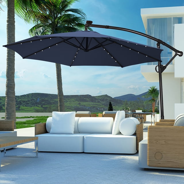 Costway 10FT Patio Offset Umbrella Solar Powered LED 360Degree Rotation Aluminum Navy. A large navy blue patio umbrella with a 10-foot canopy, featuring 24 solar-powered LED lights along its 8 steel ribs. The umbrella is supported by a powder-coated aluminum pole with a cross base for stability. It offers 360-degree rotation and tilt adjustment for optimal shade. The weather-resistant polyester fabric and vented top ensure durability and airflow. Shown in an outdoor setting with modern white and wooden furniture, surrounded by palm trees and a scenic mountain view.