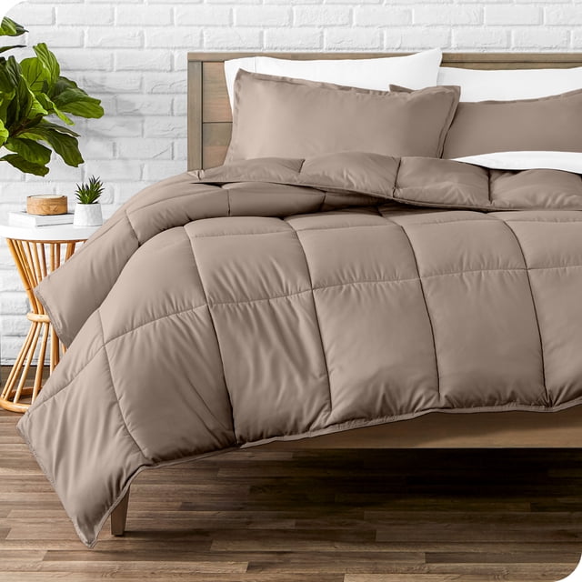 Bare Home Goose Down Alternative Comforter Set - 3 Piece Set - King/Cal King, Taupe. The set features a taupe comforter with box stitching for durability and two matching pillow shams. Crafted from hypoallergenic, ultra-soft brushed microfiber polyester, it offers breathable, all-season comfort. Shown on a wooden bed frame in a contemporary bedroom with a white brick wall and a side table with a plant.