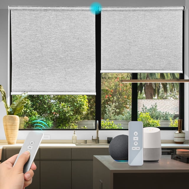 Biltek Smart Blinds for Windows, light-filtering motorized roller blinds in gray, measuring 35 inches by 72 inches. The blinds feature a cordless design with a rechargeable battery-powered motor and are controlled via a remote or smart hub compatible with Alexa and Google Hub. The image shows the blinds partially lowered in a modern kitchen setting, with a remote control and smart home devices on the counter. Made of triple-weaved textured polyester fabric with aluminum rails, these blinds provide light filtering and automation for home or office use.