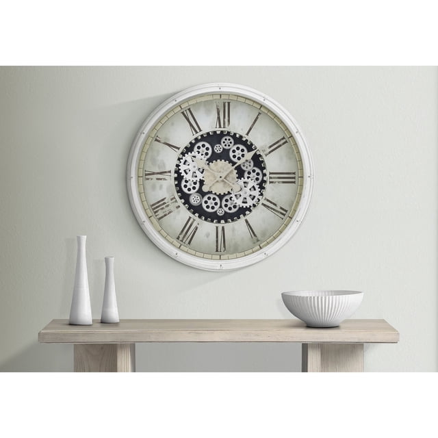 Monarch Specialties I 9033 - 30-inch round wall clock with a white plastic frame and exposed gear mechanism. Features black Roman numerals on a vintage-style face, protected by a glass surface. The clock is mounted on a light-coloured wall above a wooden console table with decorative white vases and a bowl.