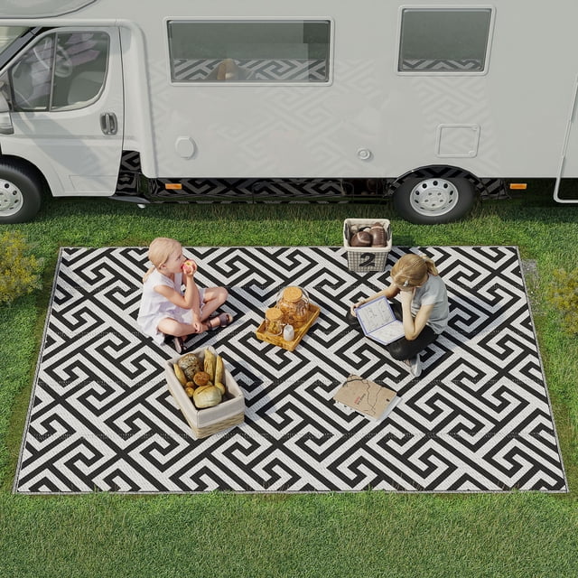 Outsunny Reversible Outdoor RV Rug, Black & White, 6' x 9', displayed outdoors near a camper van. The rug features a bold geometric black and white pattern and is set on grass with picnic items placed on top. Ideal for camping, picnics, road trips, and outdoor areas, it is waterproof, UV resistant, and reversible for versatile use.