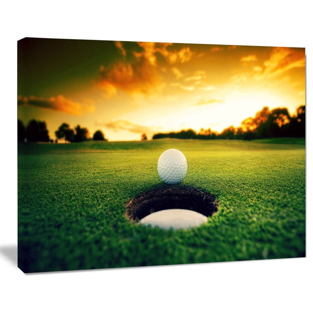 Design Art Golf Ball near Hole Landscape Artwork Canvas Print showing a close-up of a white golf ball on the edge of a golf hole on green grass with a sunset sky and tree silhouettes in the background, printed on gallery wrapped cotton canvas with a 1-inch wood subframe, ready to hang and suitable for home or office wall décor.