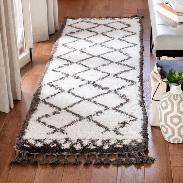 SAFAVIEH Carmella Stars Aztec Fringe Shag Area Rug with plush synthetic yarns, featuring a white base with dark gray zigzag and diamond patterns, dark gray border, and tassel fringe on the ends, placed on hardwood flooring near beige curtains and a white bench with books and a decorative vase, ideal for adding texture and warmth to contemporary spaces.