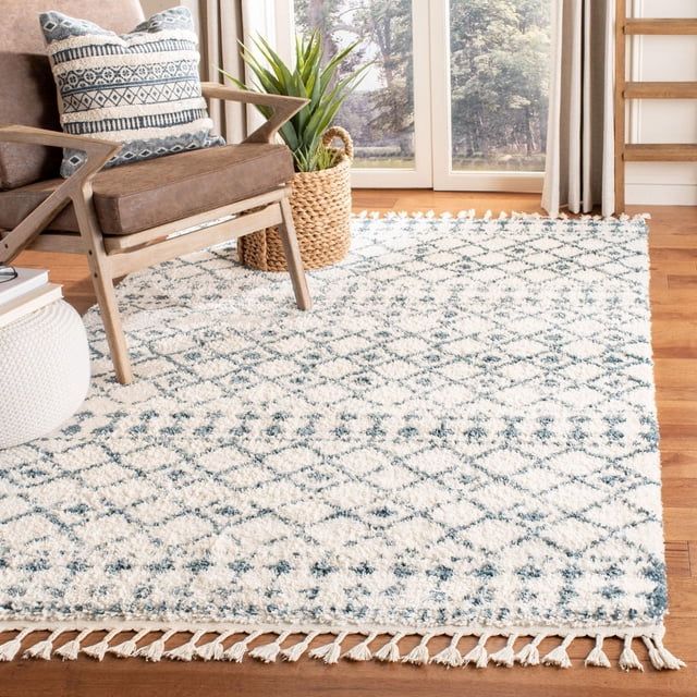 Safavieh Berber Fringe Robynne Aztec Shag Area Rug with 1.25-inch plush pile in cream and blue tribal diamond patterns, featuring fringed edges, displayed on hardwood flooring beside a wooden armchair with a patterned cushion and a woven basket plant holder, suitable for adding bohemian style to accent rug areas.