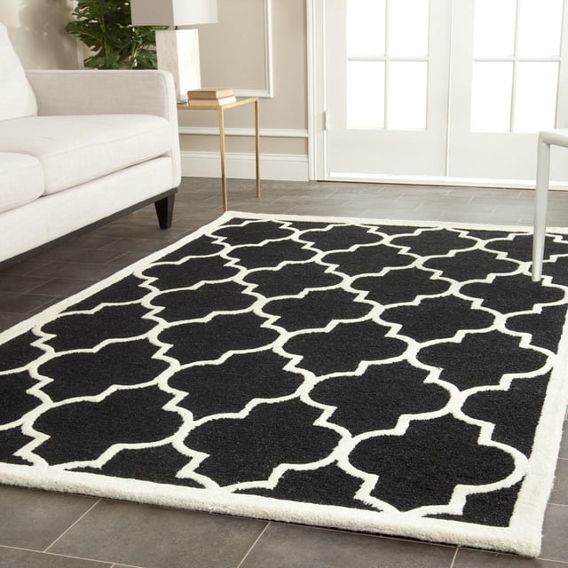 Safavieh Cambridge Kevin Geometric Area Rug with black and white geometric trellis pattern in hand-tufted wool, placed on dark tile flooring in a modern living room with beige sofa and gold side table, ideal for adding classic American style to home decor.
