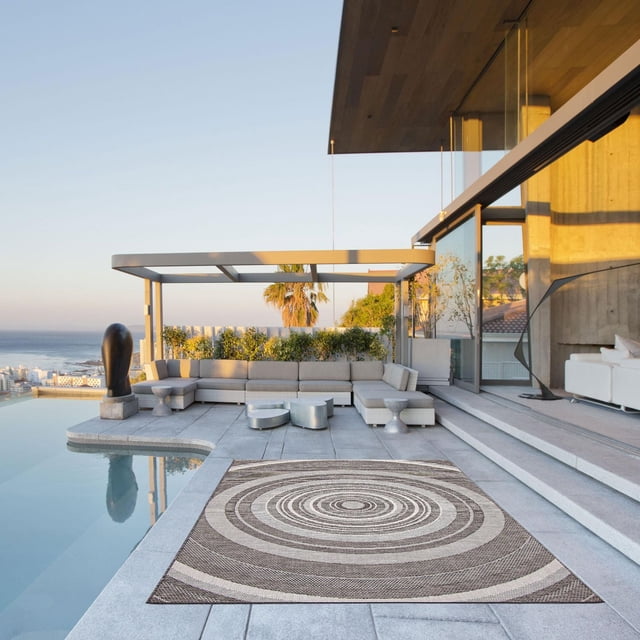 ECARPET Outdoor Area Rug for Patio, Deck, Backyard, Camping, Water Proof Carpet, Veranda Modern Collection in grey featuring a concentric circle pattern, placed on a stone patio next to an infinity pool with a modern outdoor sectional sofa under a pergola and ocean views, ideal for stylish outdoor living areas.
