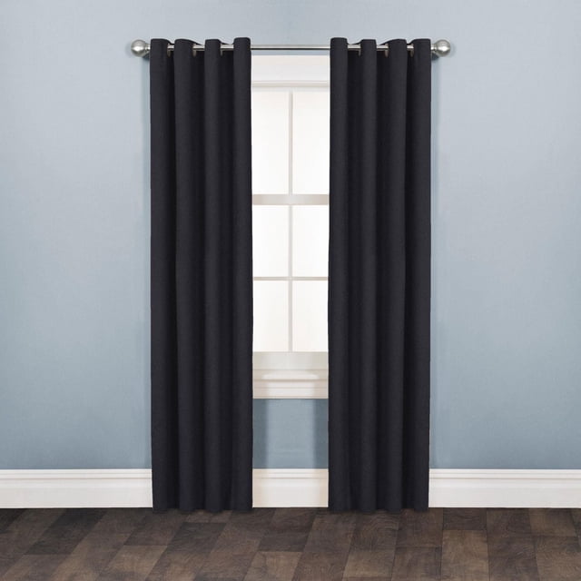 Mainstays Faux Suede Drapery Grommet Window Curtains Panel in matte black, made of soft-touch polyester with a rich suede texture, hanging on a silver curtain rod with grommet rings over a white-trimmed window, set against light blue walls and dark wood flooring, ideal for room darkening and easy machine washing.