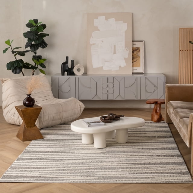 Rug Branch Hogar Abstract Indoor Outdoor Area Rug in grey and cream with a modern striped pattern, measuring 3'11" x 5'7". Made of polypropylene with jute backing, it is shown in a living room setting with a beige cushioned lounge chair, geometric wooden side table with a dark vase, white sculptural coffee table, and a grey wall-mounted cabinet with abstract geometric designs. Suitable for living room, bedroom, dining room, or office use.